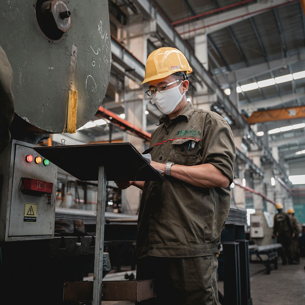 Factory worker wearing hard hat and face mask checks a clipboard beside industrial machinery