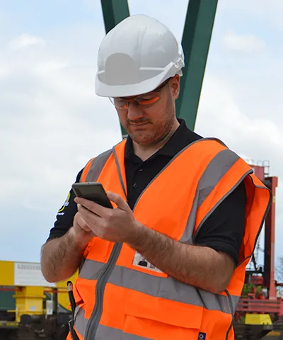 Worker in an orange safety vest and hard hat using a smartphone at a worksite