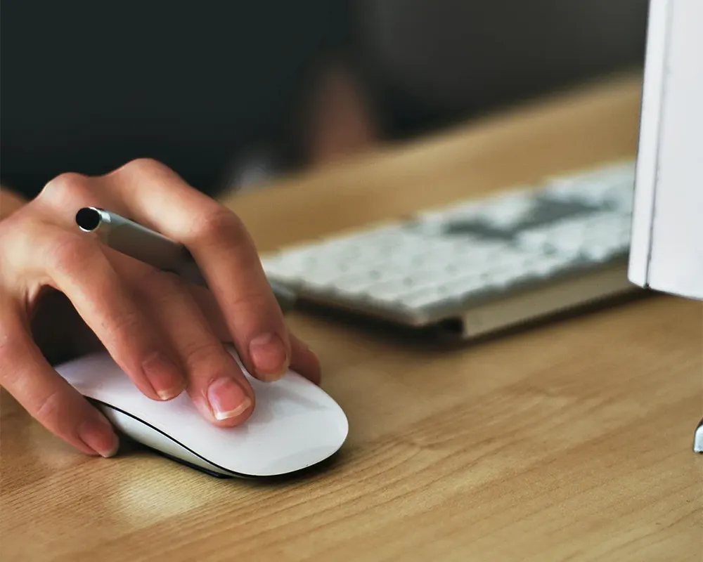 Hand using a computer mouse at a desk with a keyboard and monitor in the background