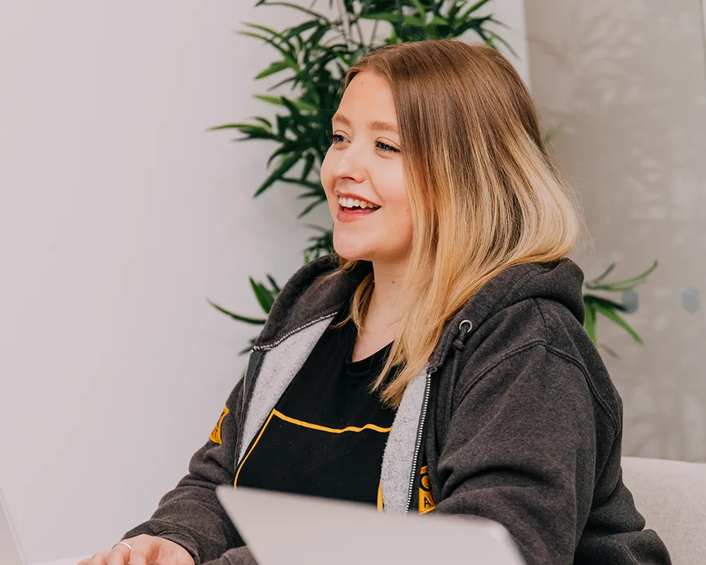 Smiling woman sitting at a desk with a laptop, wearing a hoodie in an office setting