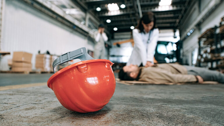 Red safety helmet on warehouse floor as worker receives CPR in the background
