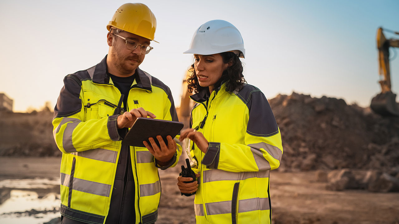 Two construction workers using a tablet while discussing plans at a job site