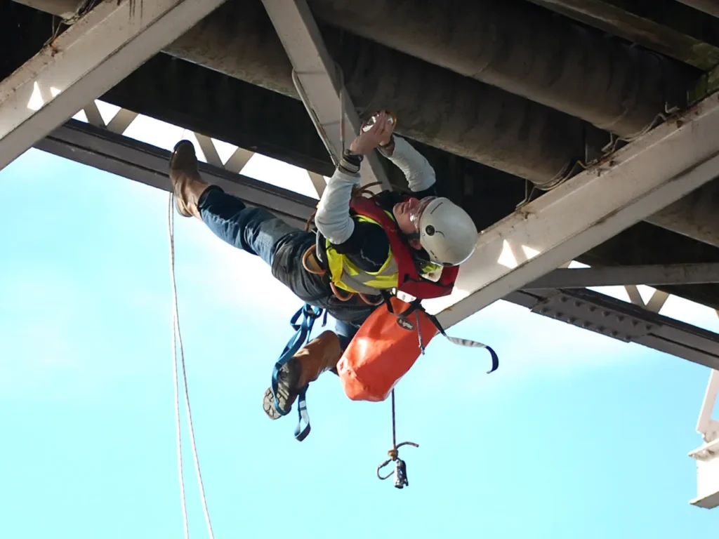 Worker suspended on ropes performing maintenance under a metal structure