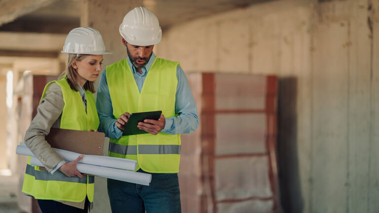 Two construction professionals reviewing plans on a tablet inside an unfinished building