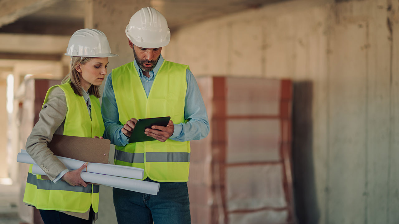 Two construction professionals reviewing plans on a tablet inside an unfinished building