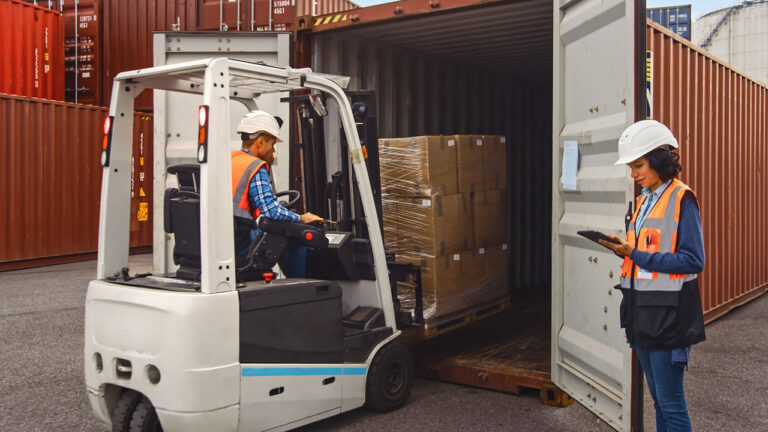 Forklift operator loading cargo container as supervisor checks tablet