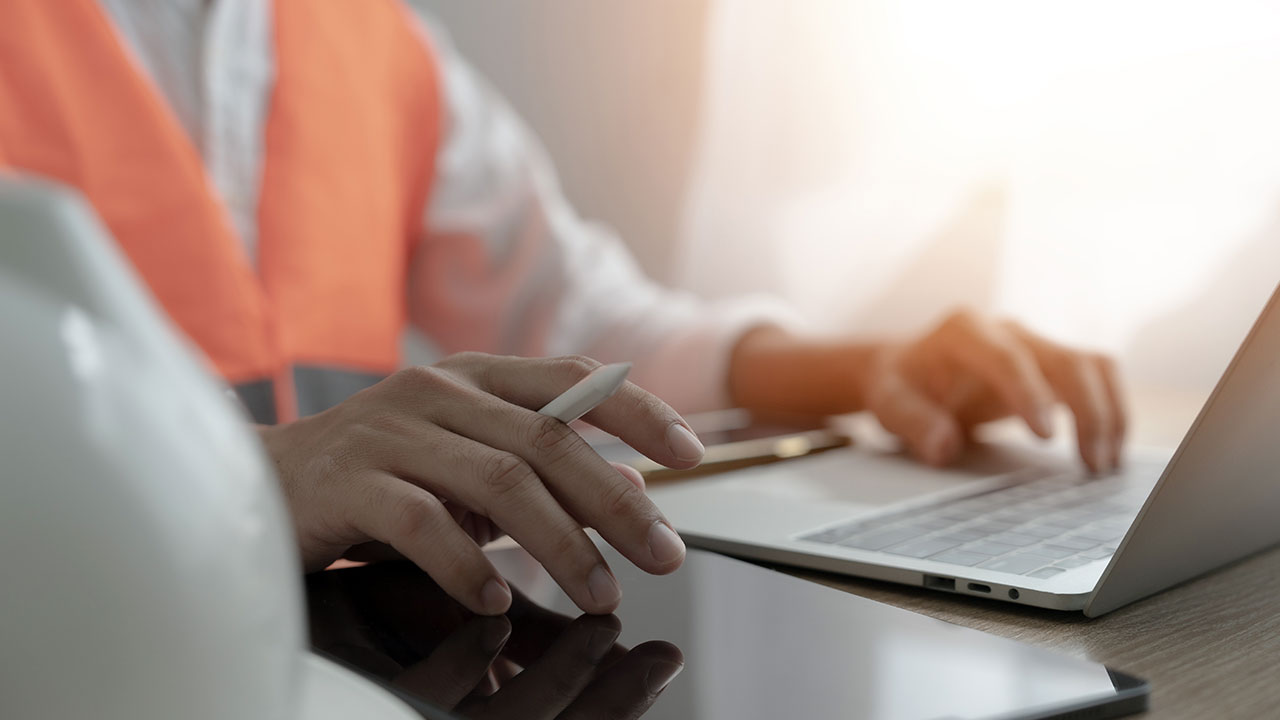 Engineer in orange vest working on laptop with hard hat nearby