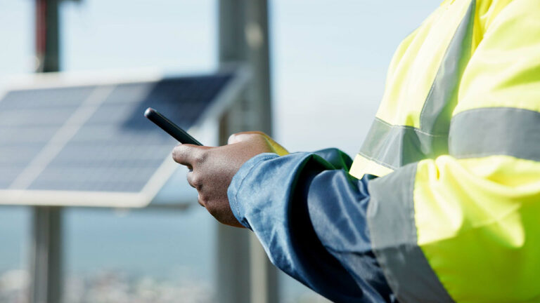Technician in reflective jacket using smartphone near solar panels