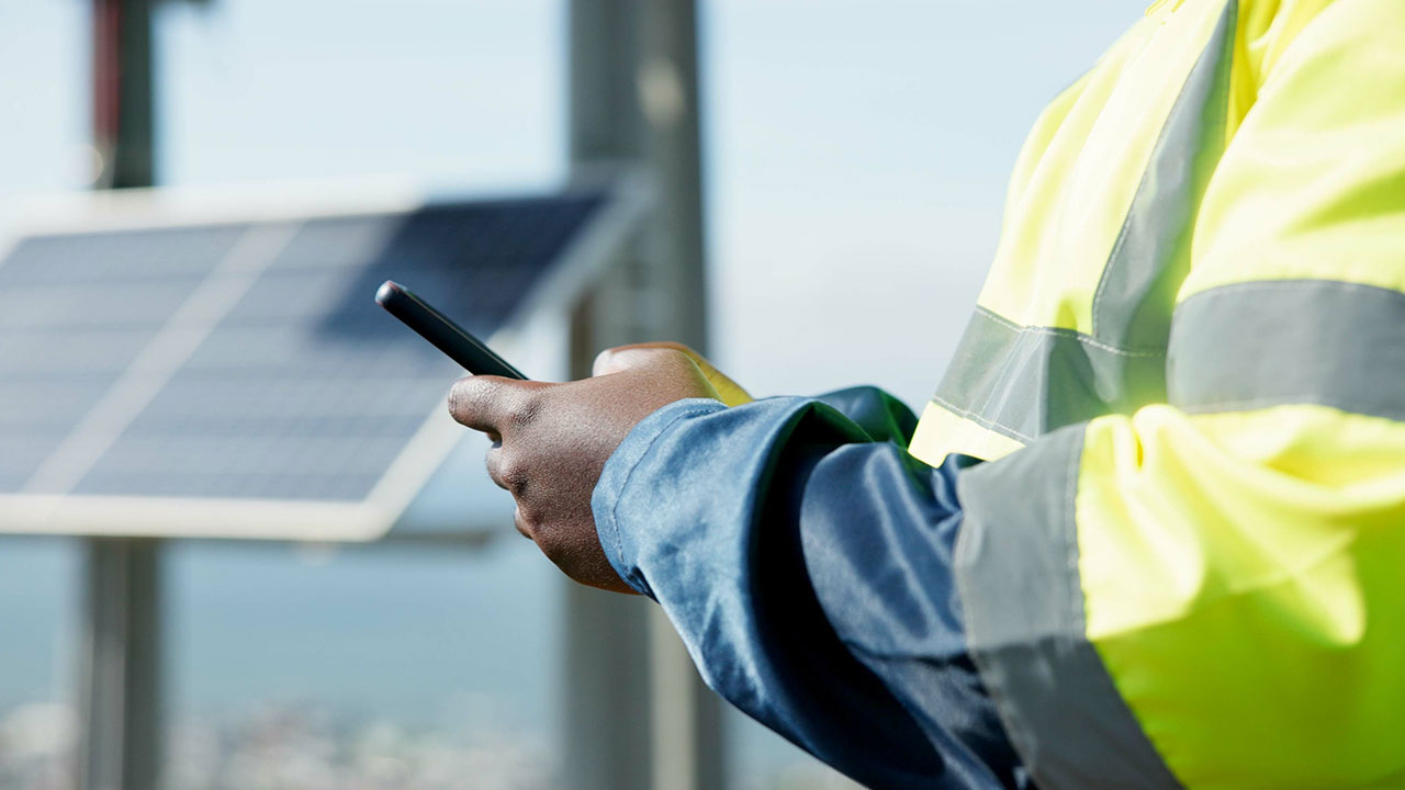 Technician in reflective jacket using smartphone near solar panels