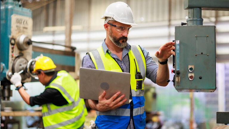 Factory worker operating industrial machinery while referencing a laptop