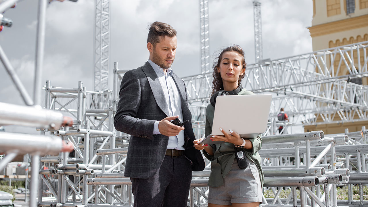 Two professionals outdoors reviewing a laptop screen near metal scaffolding