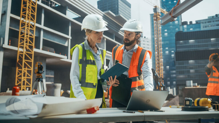 Two construction managers wearing safety vests and helmets review plans at a building site