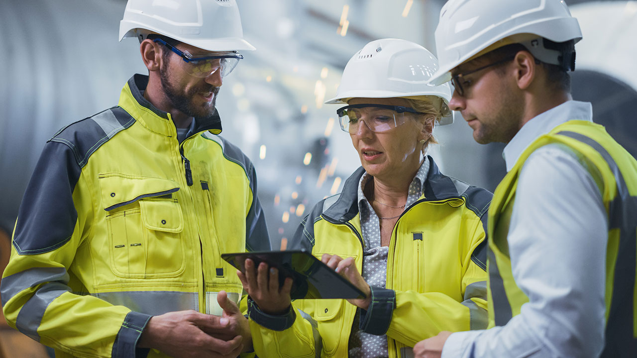 Three engineers in safety jackets and helmets discuss work while viewing a tablet near machinery