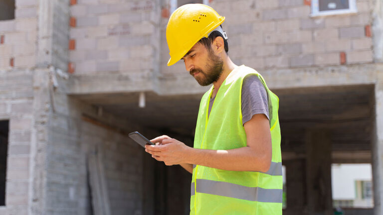 Construction worker in yellow hard hat and vest using a smartphone on-site