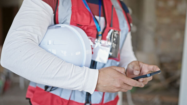 Worker in safety vest holding a hard hat and using a smartphone