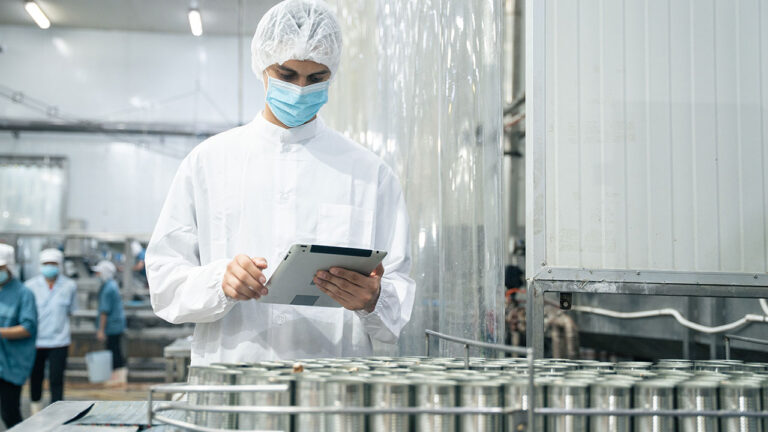 Factory worker in white uniform and mask inspecting products on tablet