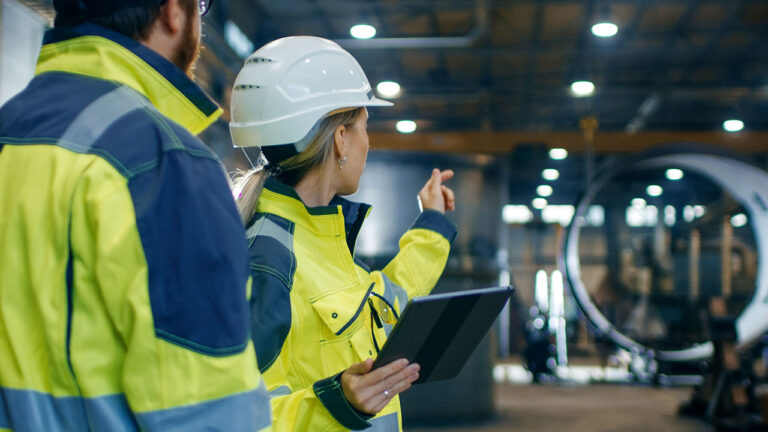 Two engineers in high-visibility jackets reviewing factory equipment with a tablet