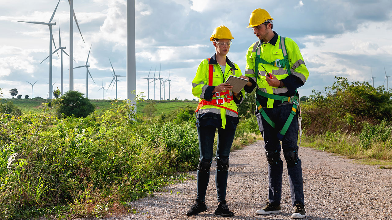 Two engineers inspecting wind turbines while reviewing data on a tablet