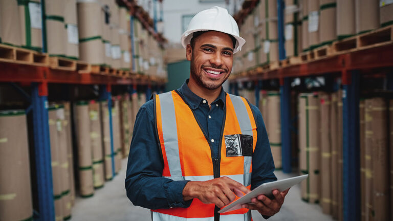 A smiling warehouse worker in a hard hat and safety vest uses a tablet among stacked goods
