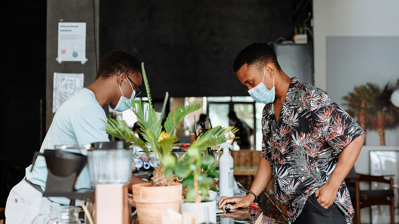 Two people wearing face masks interact at a counter inside a cafe or workplace