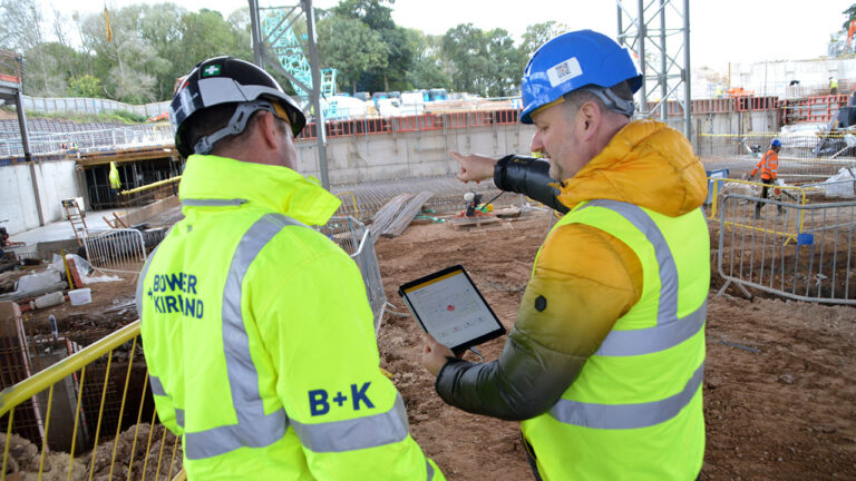 Two construction workers review a tablet on site with safety gear and machinery in the background