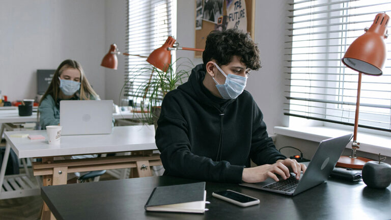 Two office workers wearing face masks work on laptops at separate desks