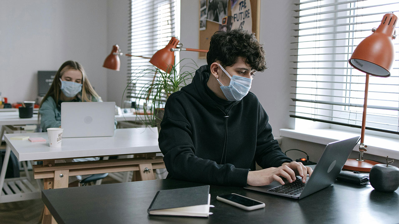Two office workers wearing face masks work on laptops at separate desks