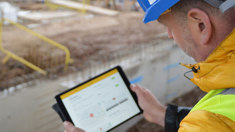 A construction worker uses a tablet on site while wearing a blue hard hat and yellow jacket