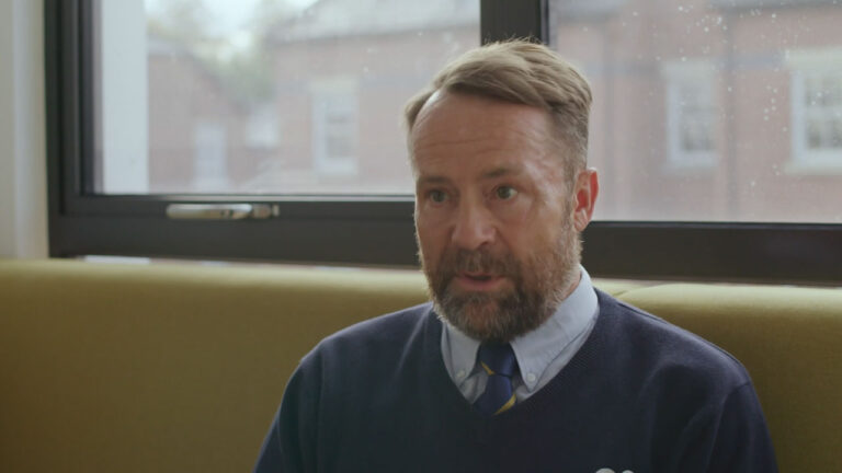 A man in a blue sweater and tie speaks while seated indoors