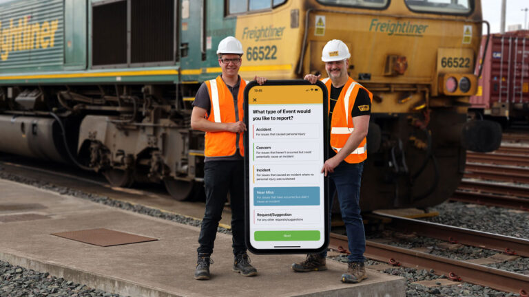 Rail workers in safety vests demonstrate a mobile safety reporting app beside a train