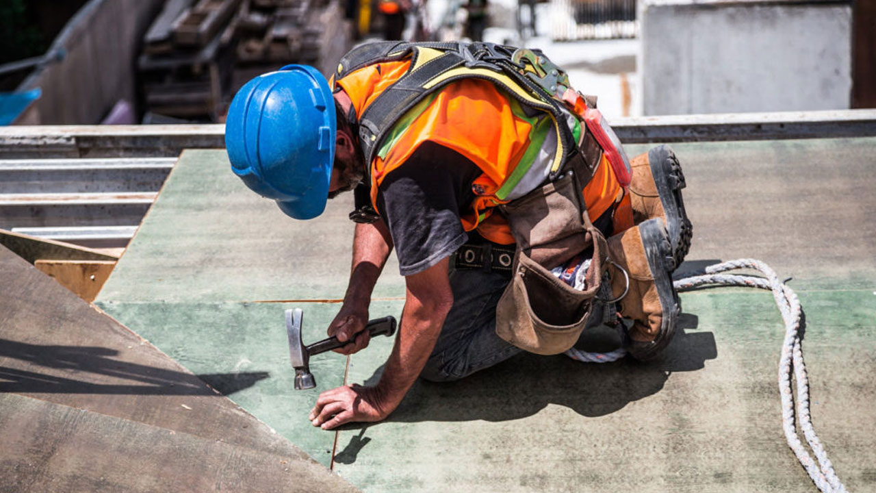A construction worker in safety gear hammers nails while secured by a harness on site