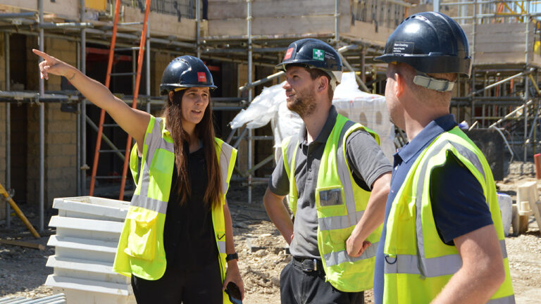 Three construction workers in safety gear discuss plans on site as one points ahead