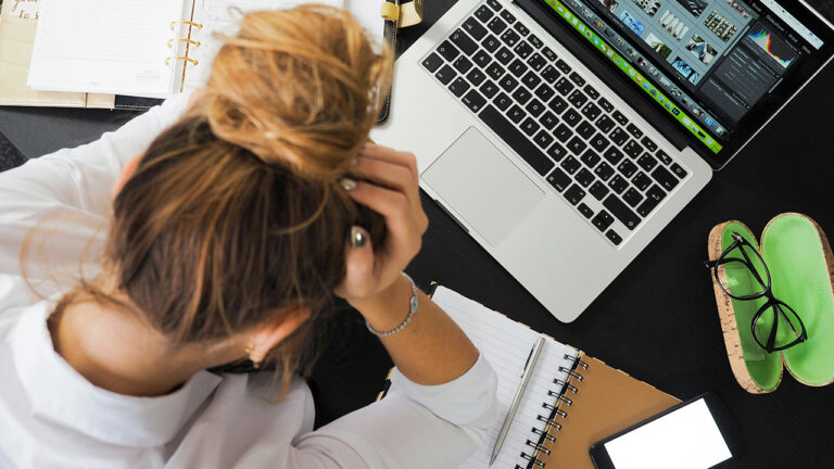 A stressed office worker sits at a desk holding their head beside a laptop and notebooks