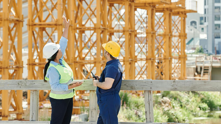 Two construction workers wearing safety gear discuss and point toward scaffolding on site