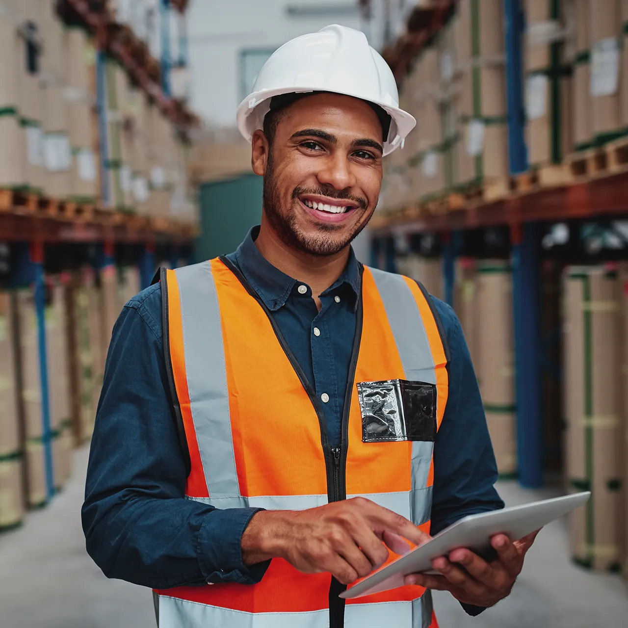 A smiling warehouse worker in a hard hat and safety vest uses a tablet among stacked goods