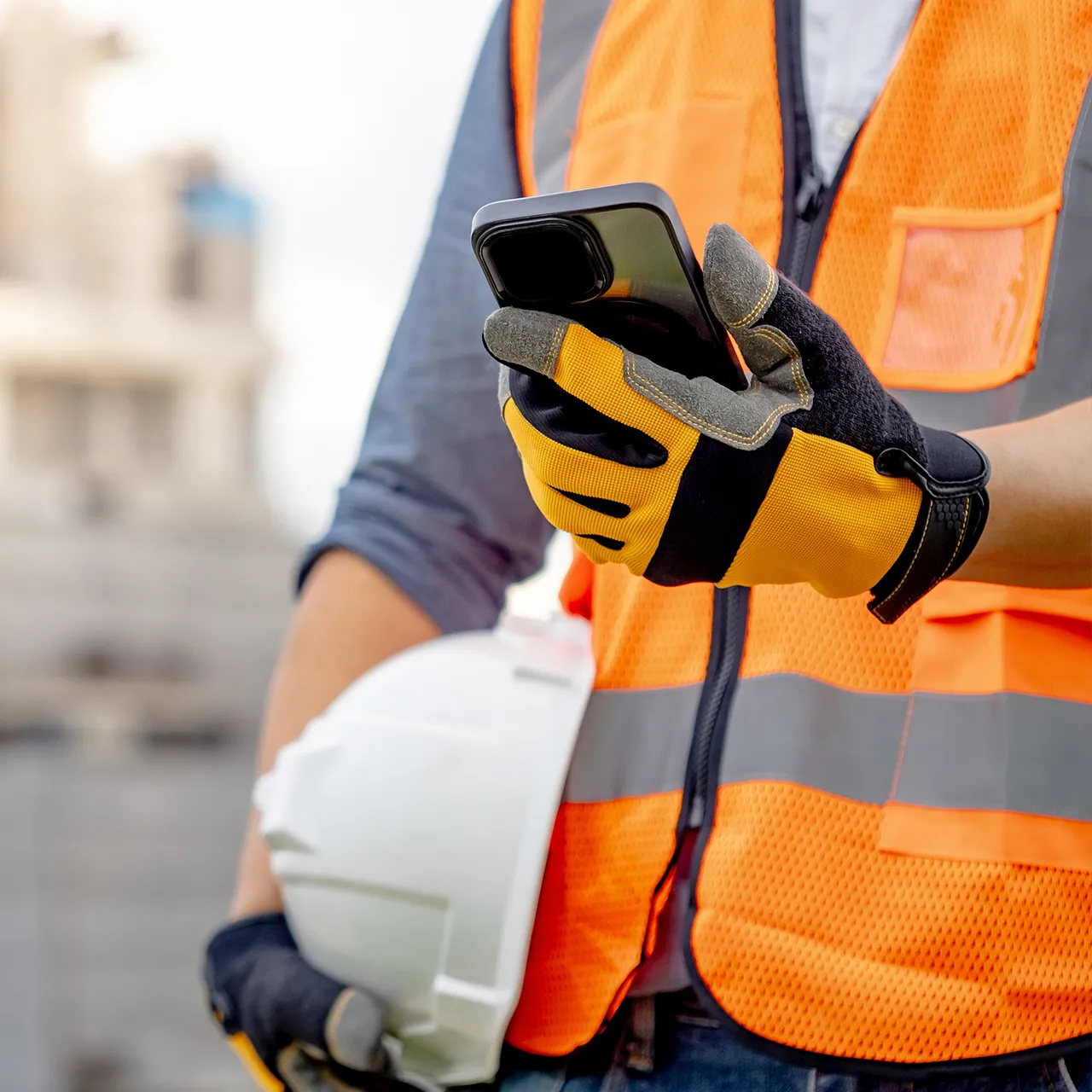 A construction worker wearing gloves and a safety vest uses a smartphone on site