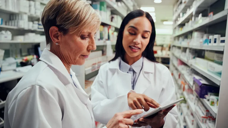 Two pharmacists in lab coats review a tablet in a pharmacy aisle