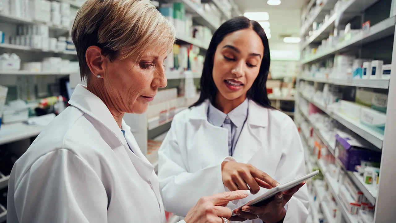 Two pharmacists in lab coats review a tablet in a pharmacy aisle