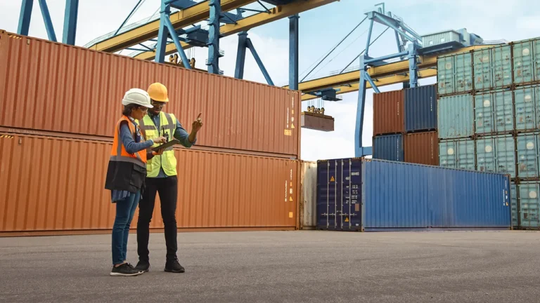 Two workers in hard hats review tablet at shipping yard with stacked containers