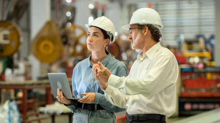 Two engineers in hard hats discuss factory operations while holding a laptop.