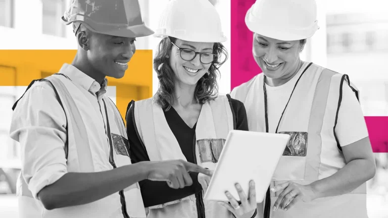 Three construction workers in hard hats and safety vests review a tablet.